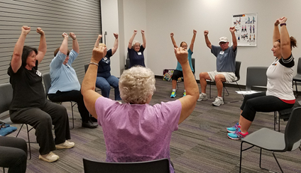 Older adults sit in chairs in a big circle, facing one another. They watch the instructors as they lift their arms above their heads.