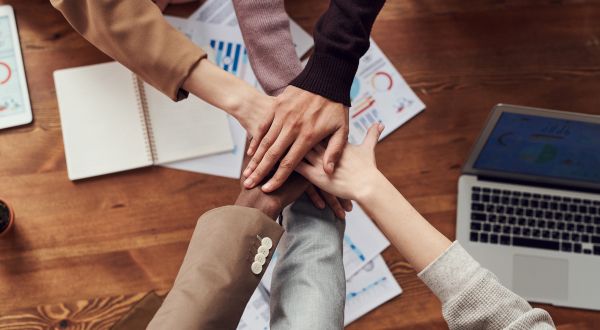 a variety of 5 hands on top of each other in above a table with business documents and a laptop