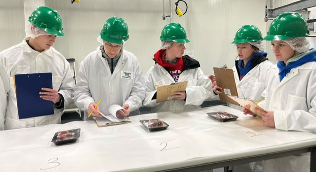 Five 4-H members wearing green hard hats standing around table